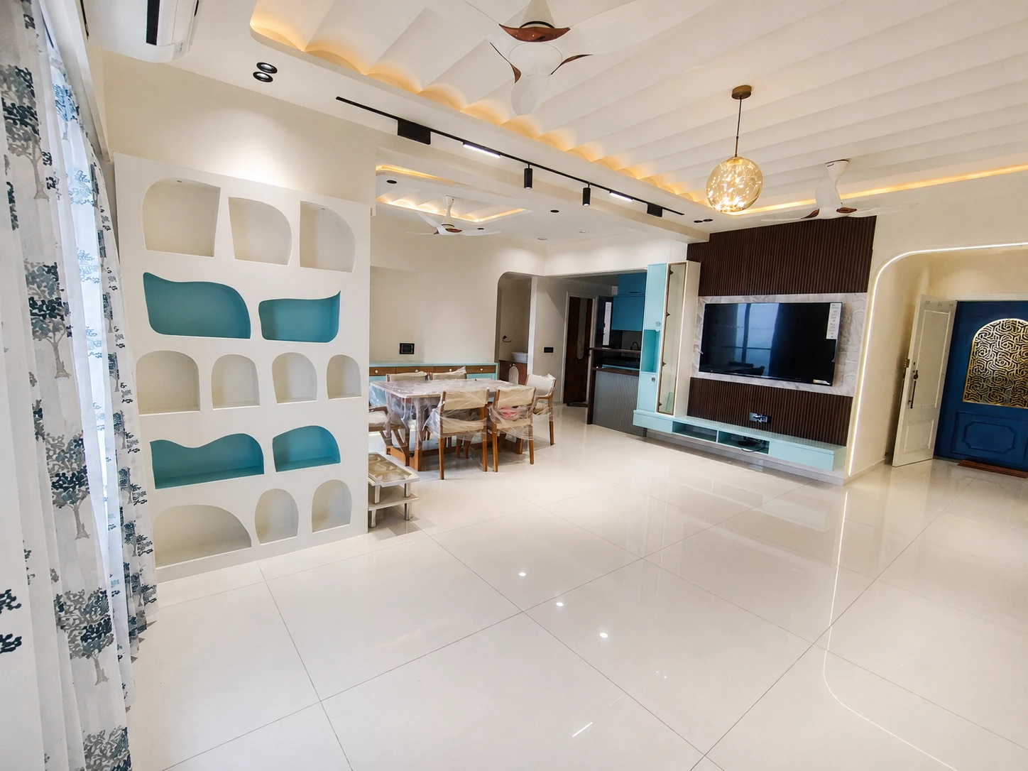 This spacious, modern living and dining area embraces a minimalist aesthetic with warm, ambient lighting and sleek design. The room features contemporary elements like glossy white tiles, wooden accents, vibrant teal tones, and geometric shelving, all set against a backdrop of clean, crisp architectural details and thoughtfully integrated lighting.